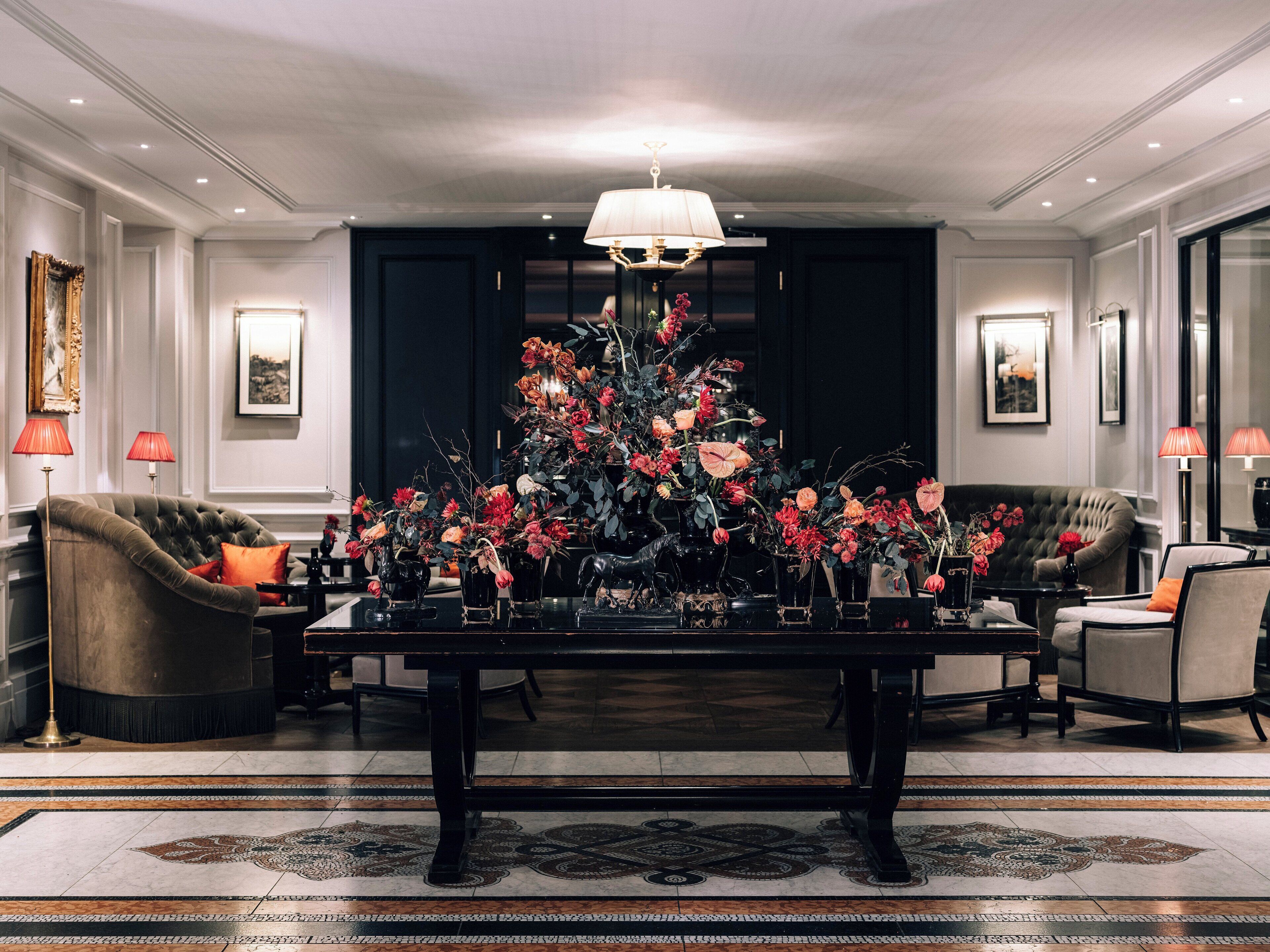 A luxurious living room with a large dark wooden coffee table surrounded by elegant chairs, a grand chandelier, and a floral centerpiece.