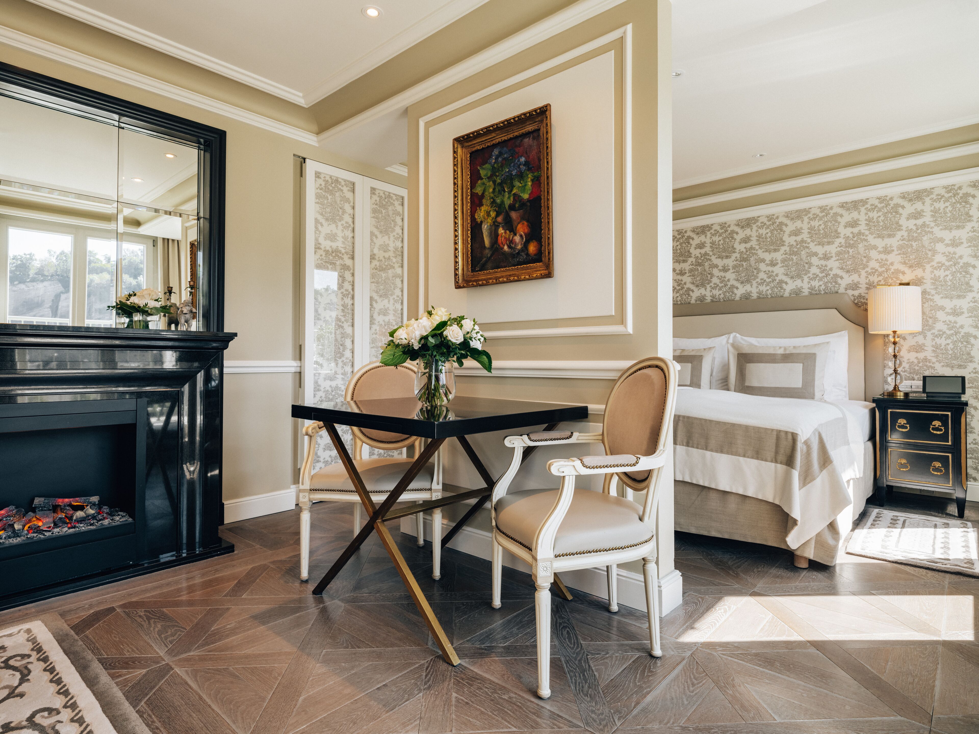 A hotel room with a bed, a desk with two chairs, a fireplace, and a painting on the wall.