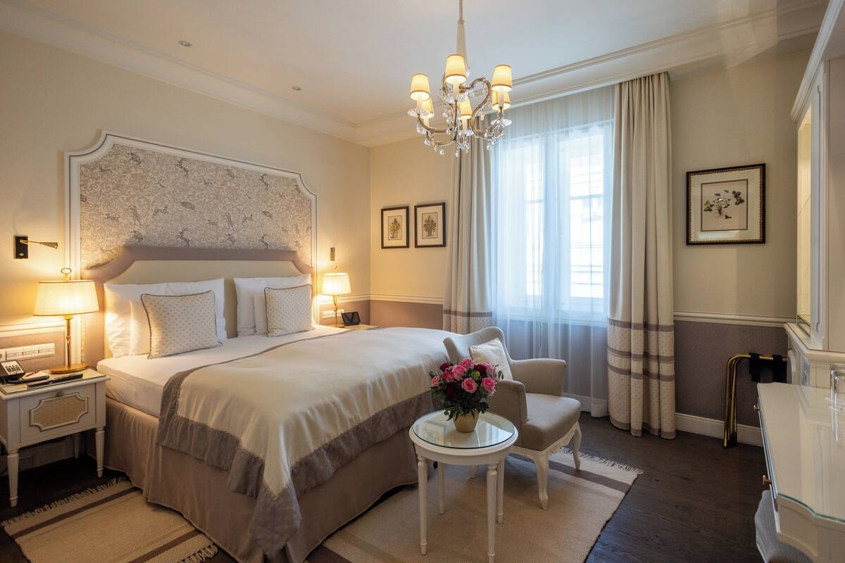 A hotel room with a large bed, bedside lamps, a small table with flowers, a chair, and a chandelier.