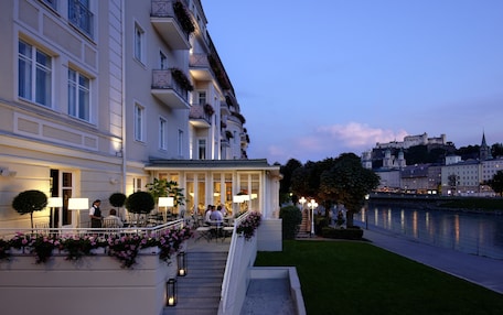 A hotel with a grand entrance, outdoor seating area, and a view of a castle across the river.