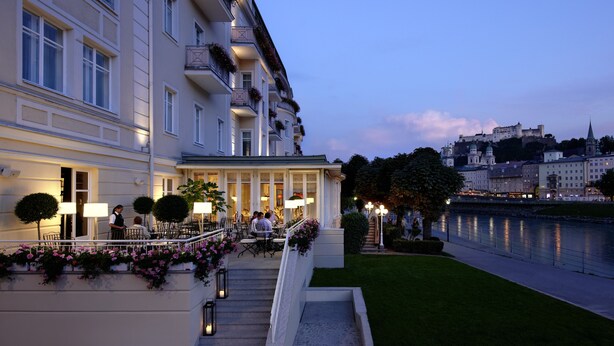 A hotel with a grand entrance, outdoor seating area, and a view of a castle across the river. - Hotel Sacher Salzburg (Salzburg)