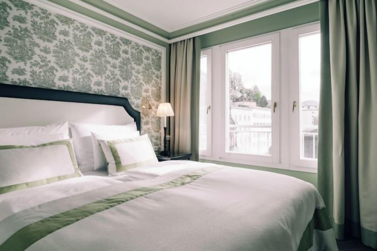 Habitación de hotel con una cama grande, una mesita de noche, una lámpara y vistas a un edificio a través de una ventana.