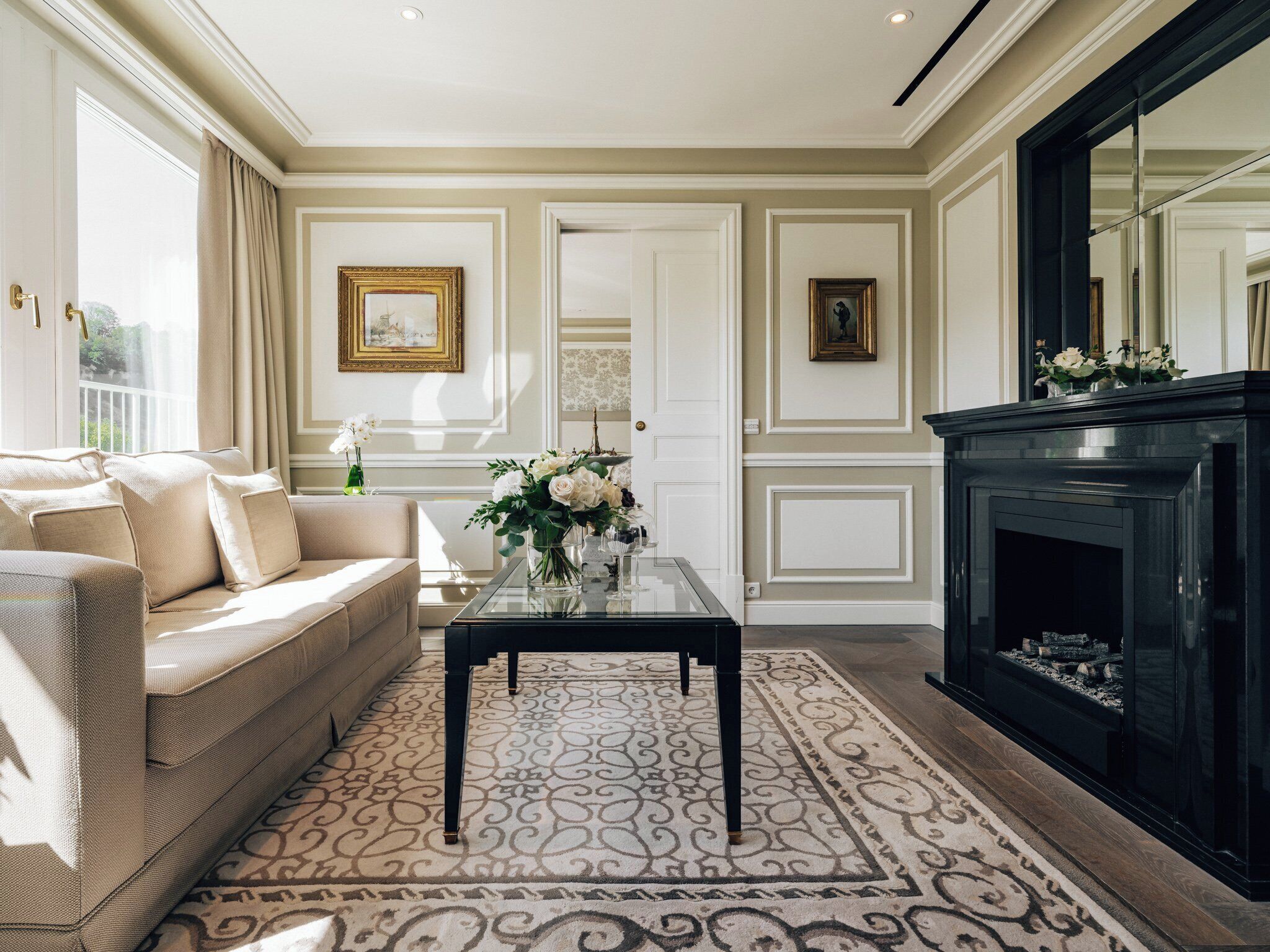 Una sala de estar con un sofá color beige, una mesa de centro de cristal con flores, una chimenea y cuadros en las paredes.