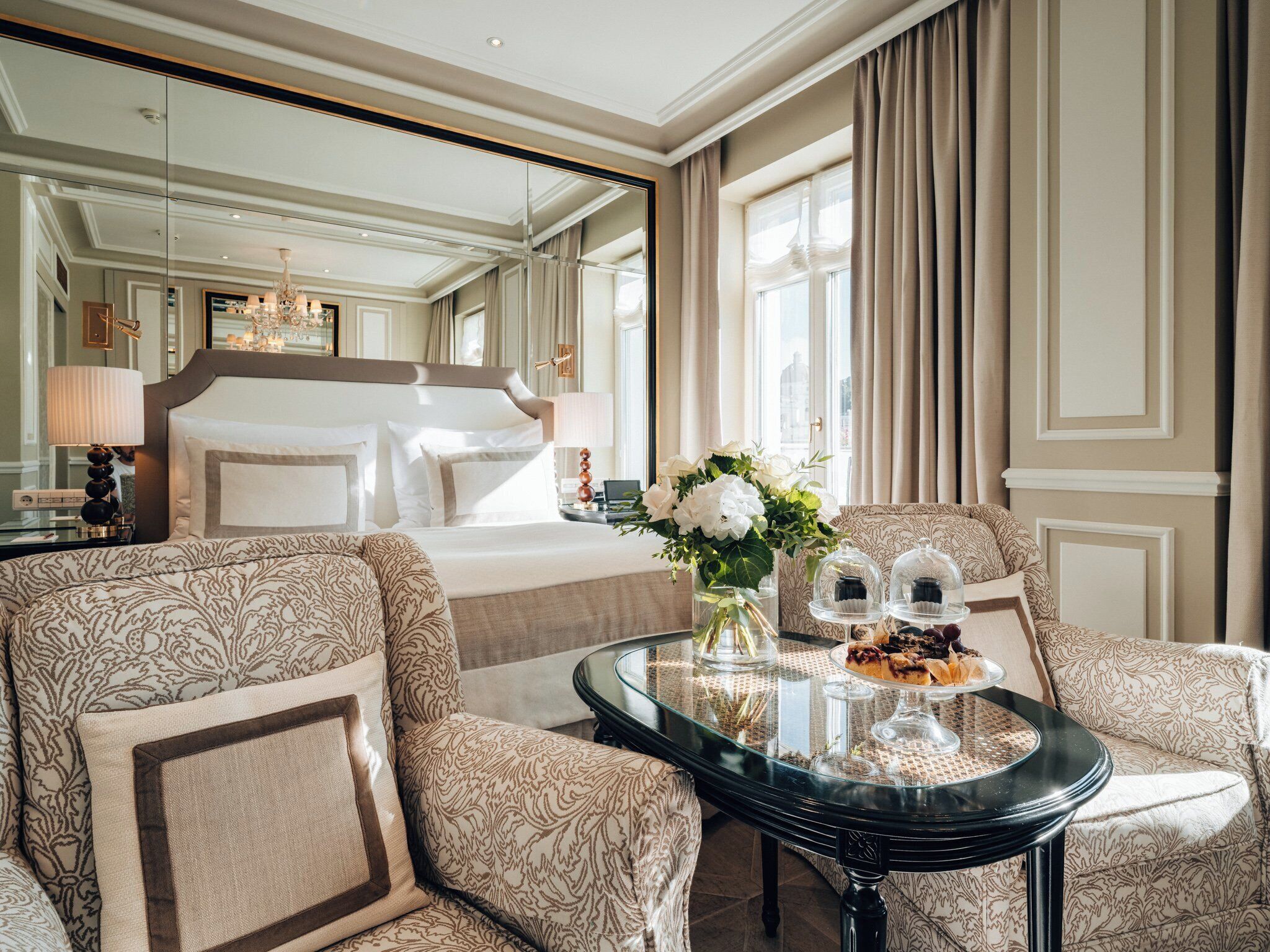 A luxurious hotel room with a large bed, a sitting area with patterned sofas, a glass coffee table, and a view of the city through large windows.