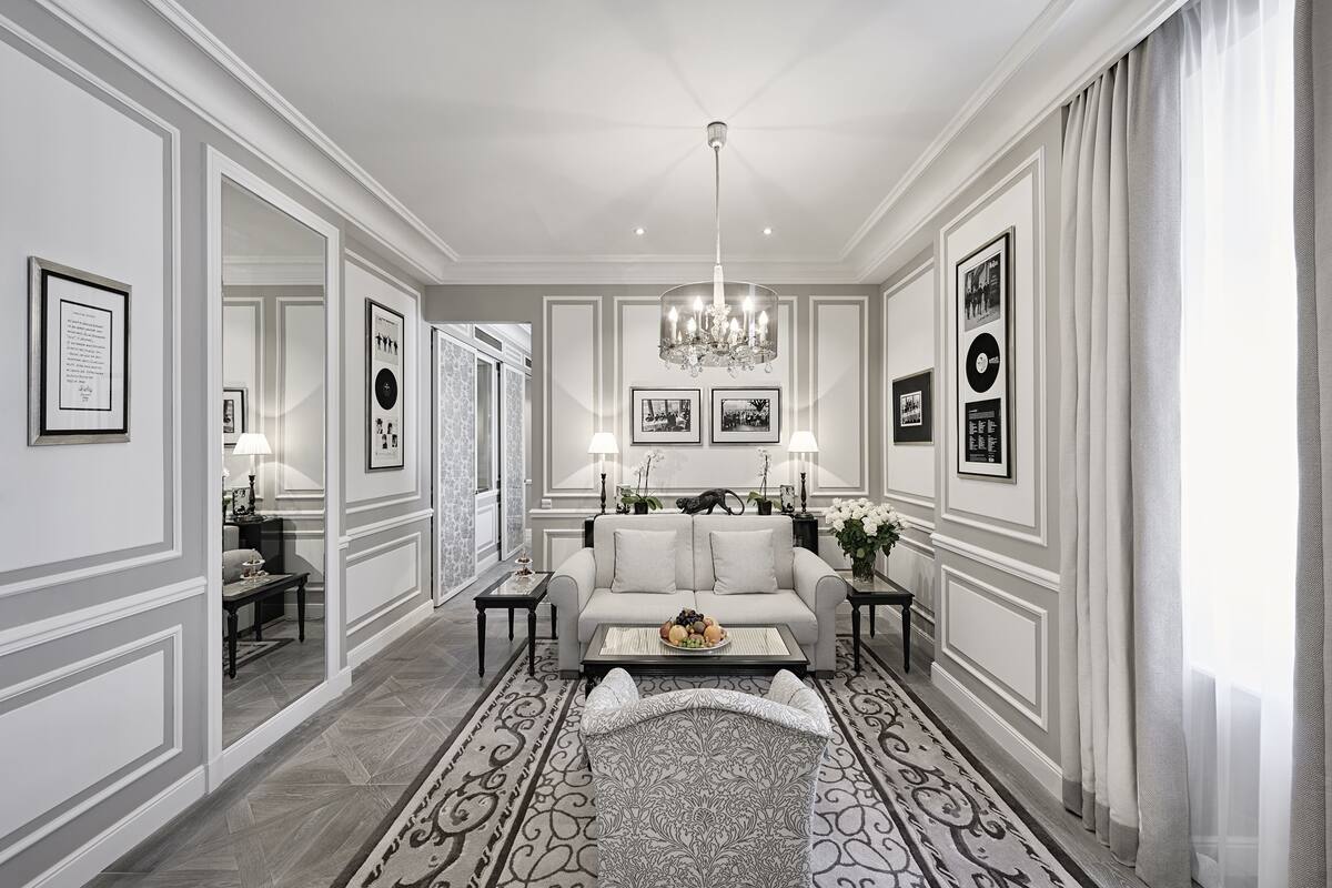 A spacious living room with a large sofa, a coffee table, and a chandelier. There are framed pictures on the walls and a patterned rug on the floor.
