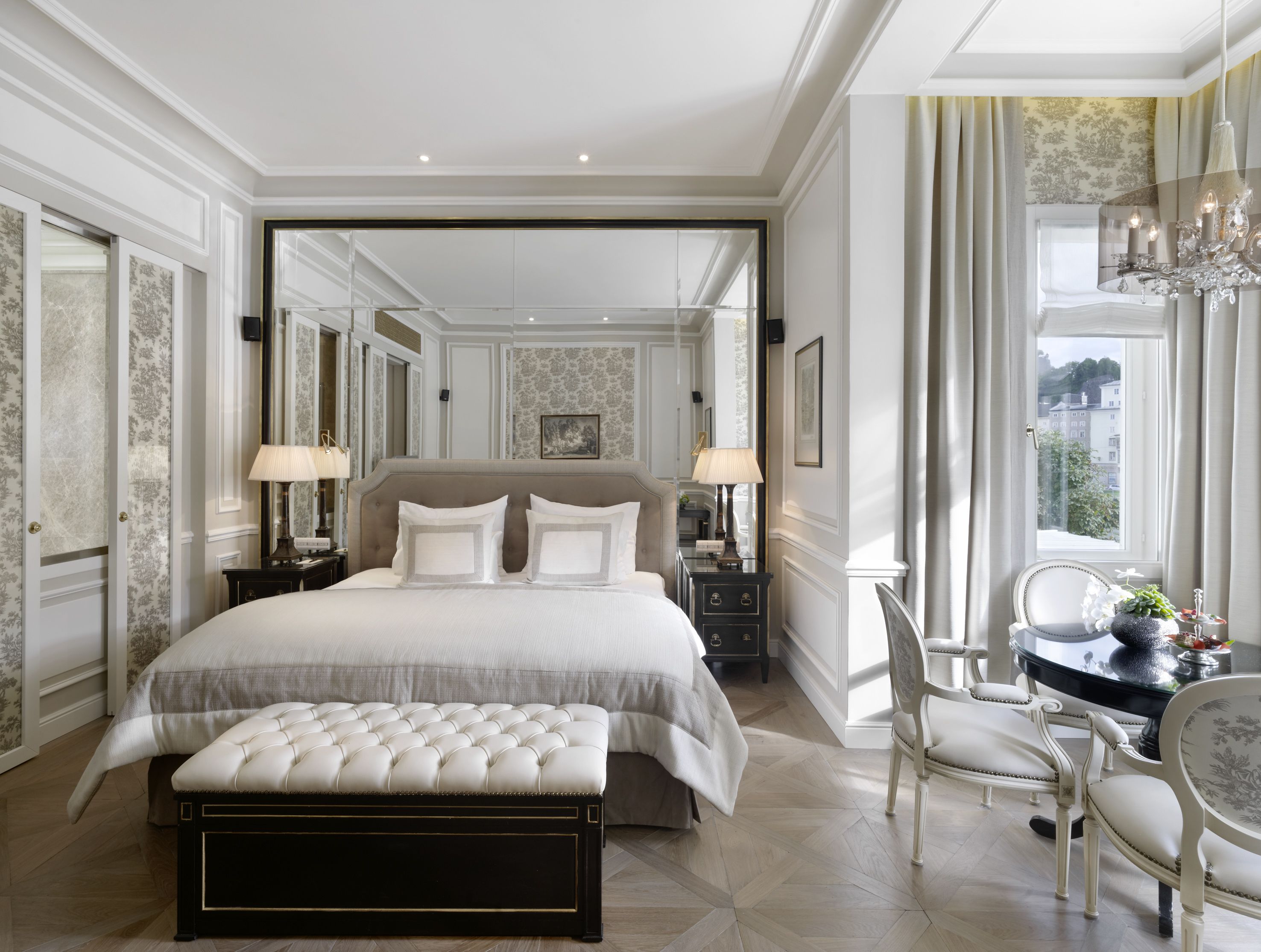 A luxurious bedroom with a large bed, a sitting area with a table and chairs, and a view of the outdoors through a window.