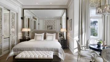 A luxurious bedroom with a large bed, a sitting area with a table and chairs, and a view of the outdoors through a window.