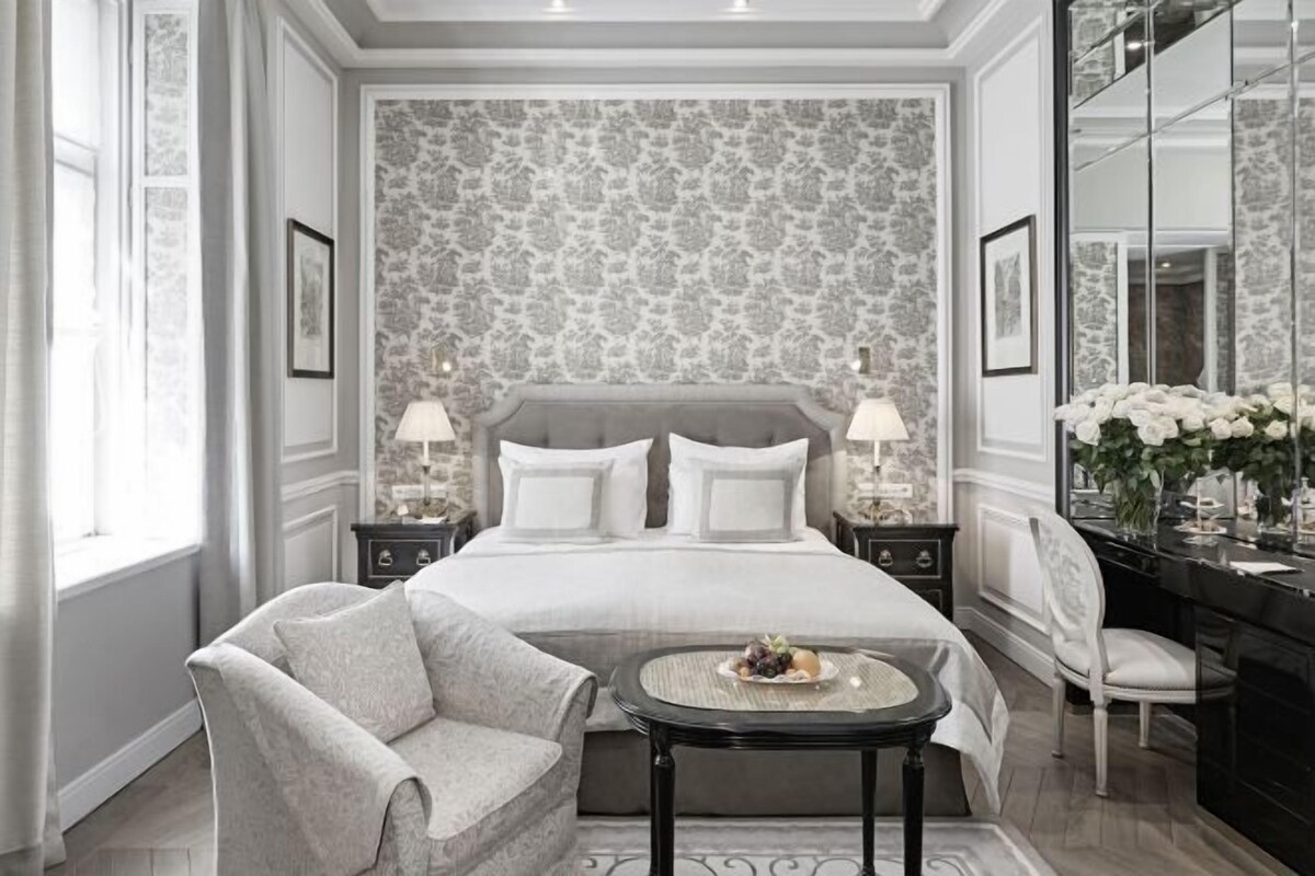 Habitación de hotel con una cama grande, dos mesitas de noche, una zona de estar con una silla y una mesa, un tocador con espejo y una ventana con cortinas.