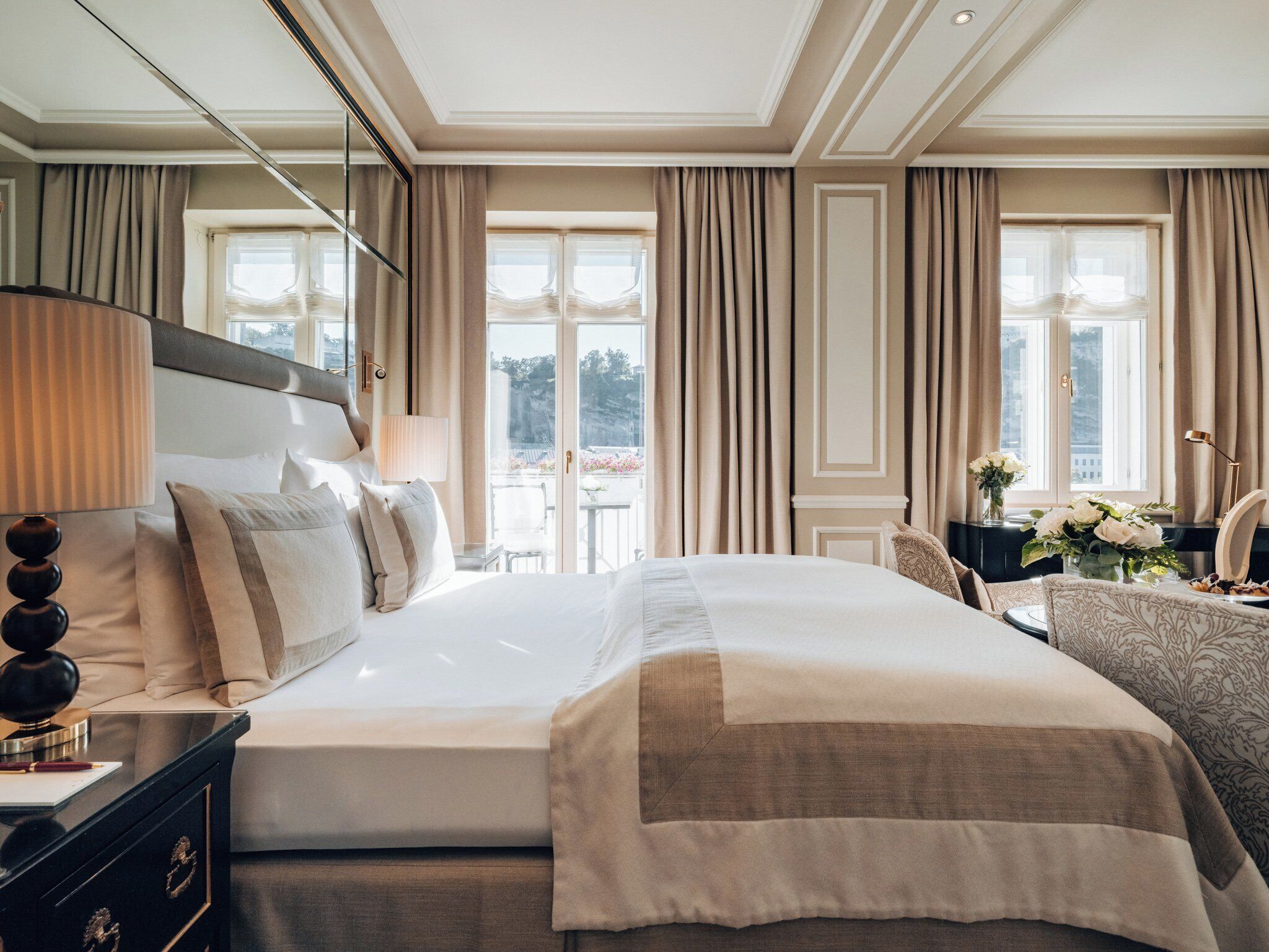 A spacious hotel room with a large bed, a bedside table with a lamp, a dresser, and a view of the outdoors through large windows.