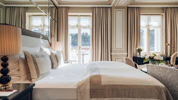 A spacious hotel room with a large bed, a bedside table with a lamp, a dresser, and a view of the outdoors through large windows.