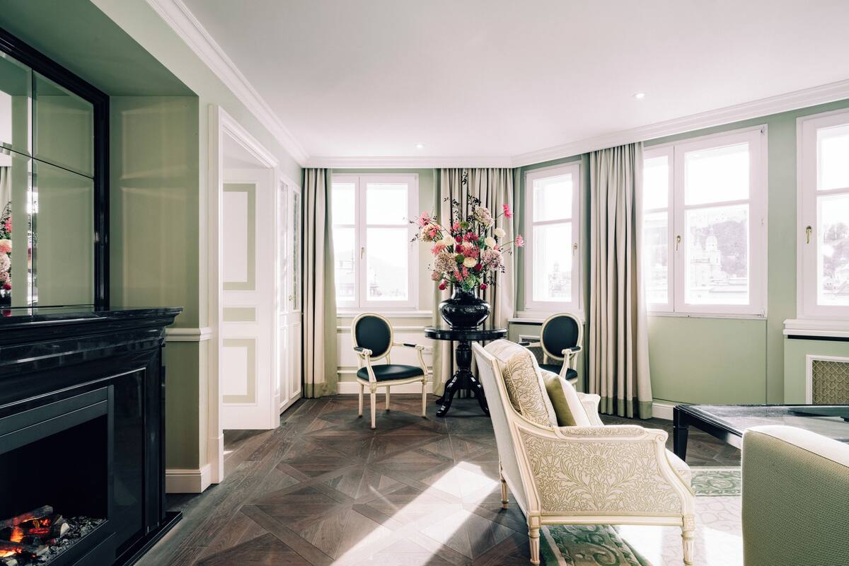 A room with a fireplace, two chairs, a table with a vase of flowers, and large windows with curtains.