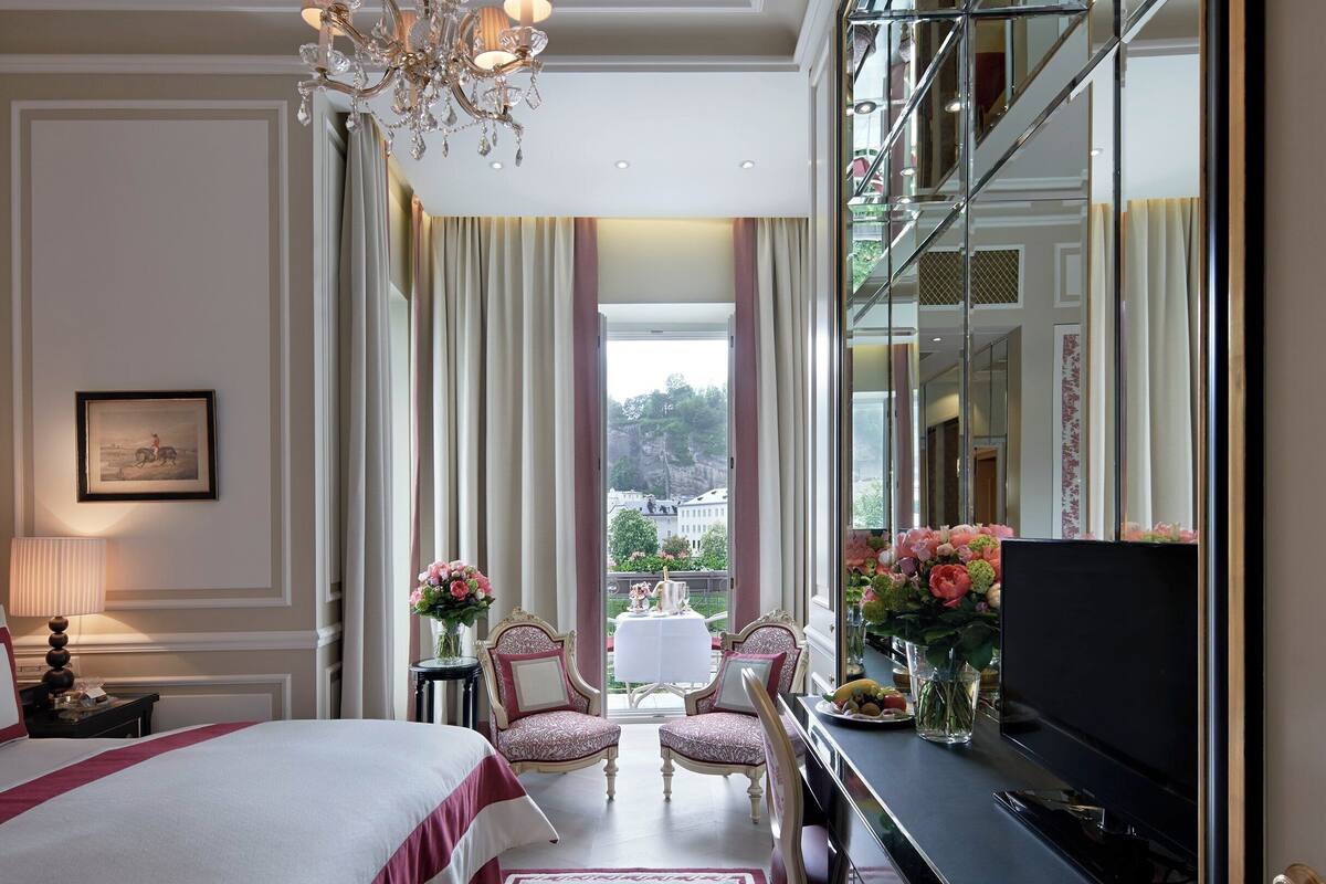 A hotel room with a large bed, a television, a chandelier, a mirror, and a view of the outdoors.