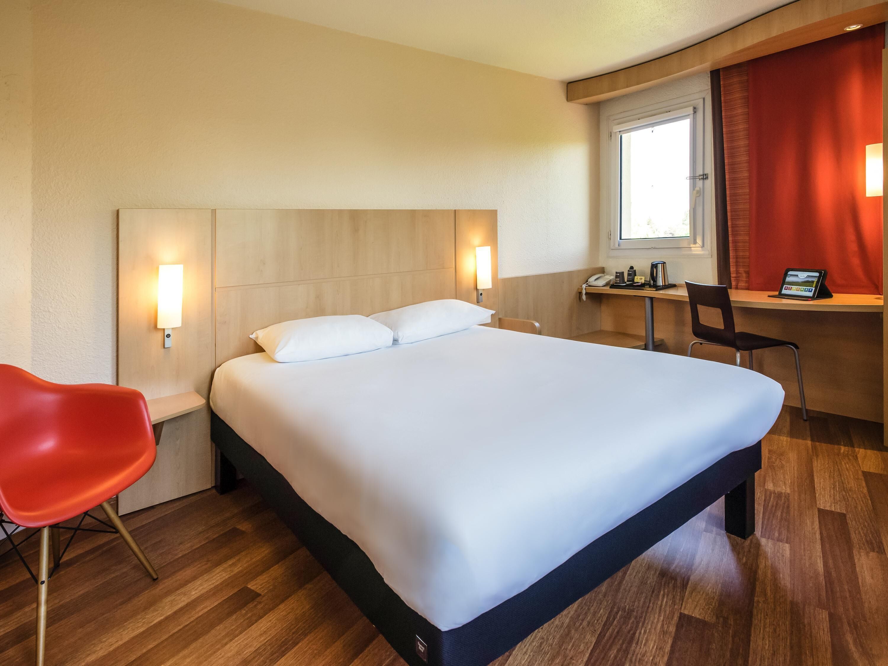 Habitación de hotel con una cama grande, dos lámparas de noche, un escritorio con una silla, una silla roja y una ventana con cortinas.