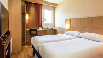 A hotel room with two beds, a desk, a TV, and a window with curtains.