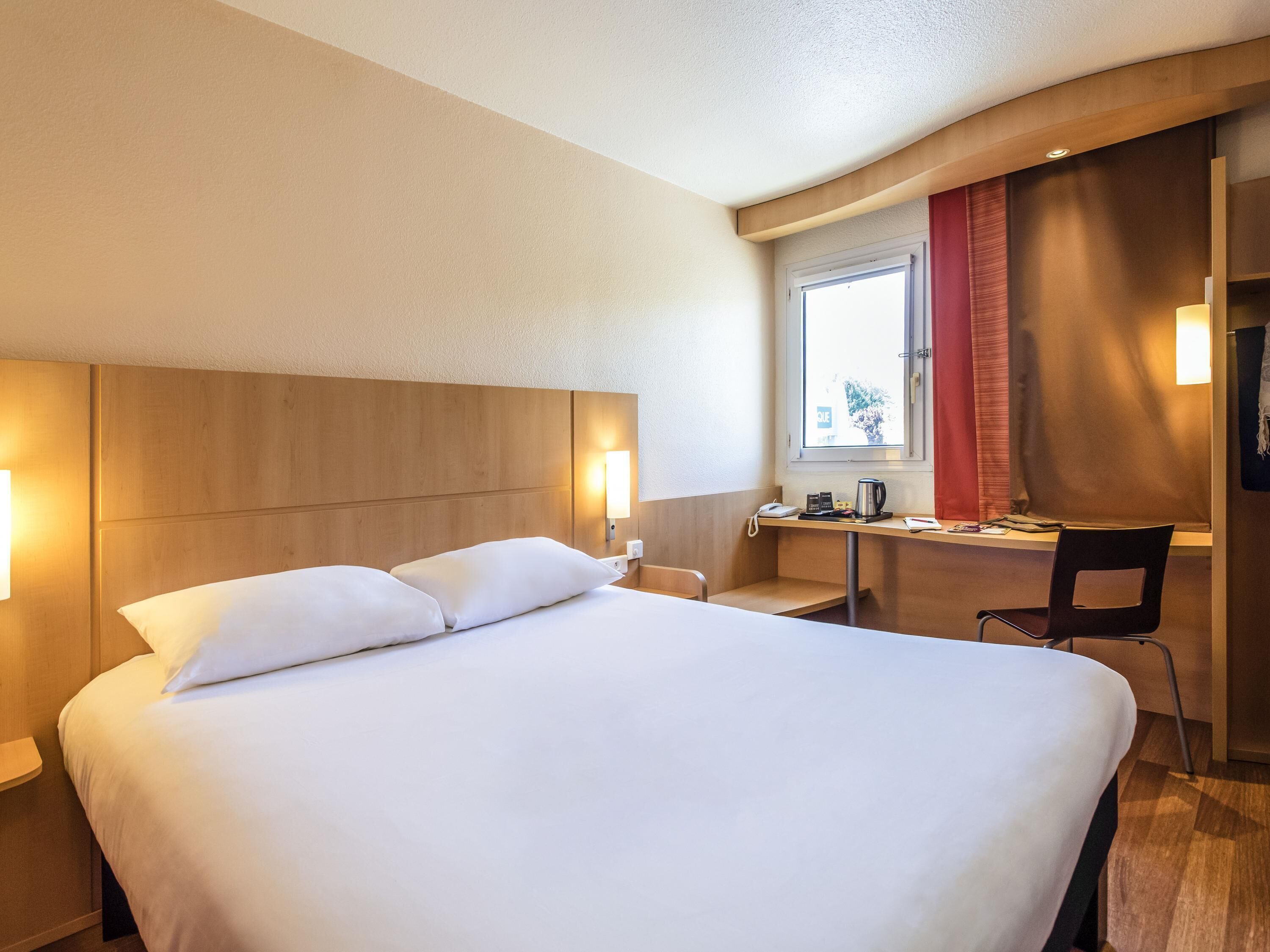 A hotel room with a large bed, a desk with a chair, a window, and a door.
