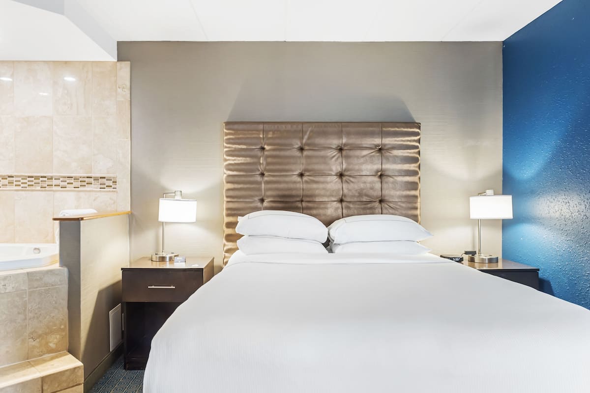 A hotel room with a large bed, two bedside tables, a nightstand, a wall-mounted shelf, and a bathroom with a bathtub.