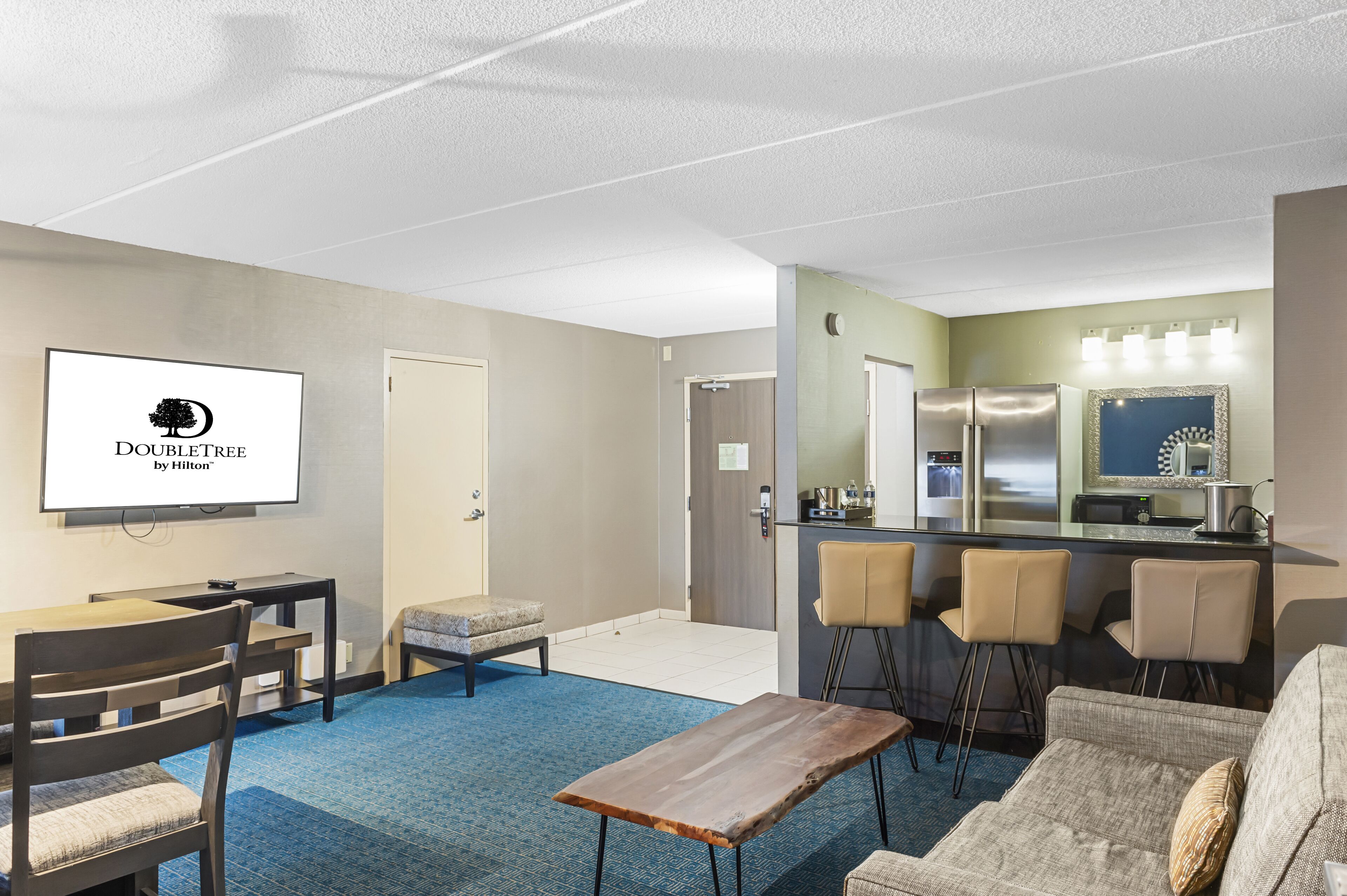 A hotel room with a TV displaying "DOUBLETREE by Hilton", a kitchenette, a seating area with a sofa and chairs, and a blue carpet.