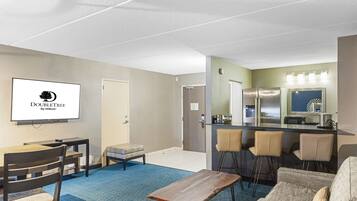 A hotel room with a TV displaying "DOUBLETREE by Hilton", a kitchenette, a seating area with a sofa and chairs, and a blue carpet.