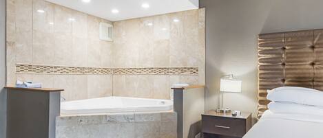 A hotel room with a large bathtub, a bed, a nightstand with a lamp, and a mirror on the wall.