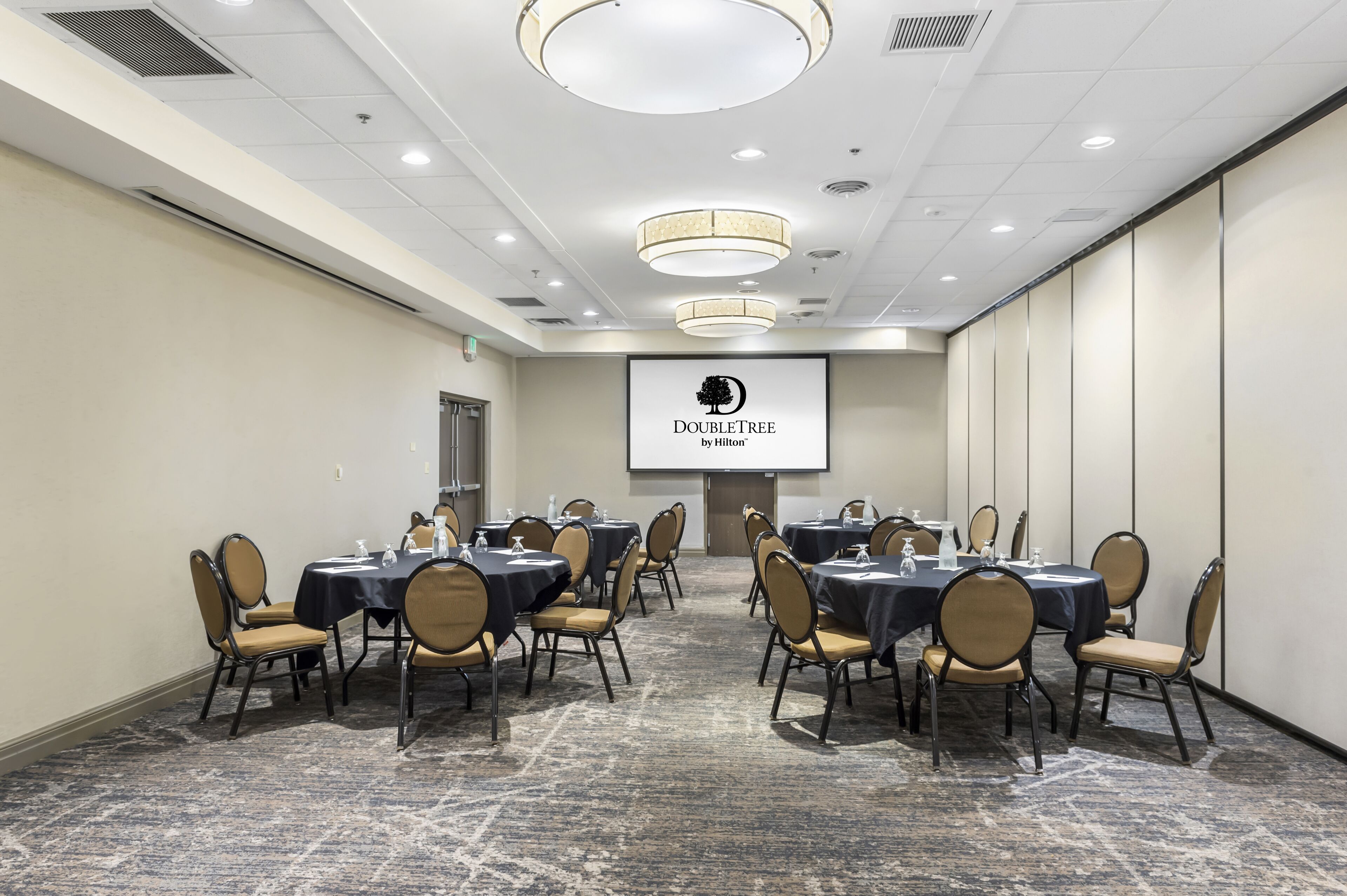 A conference room with multiple tables and chairs, a projector screen displaying "DOUBLETREE by Hilton", and a carpeted floor.