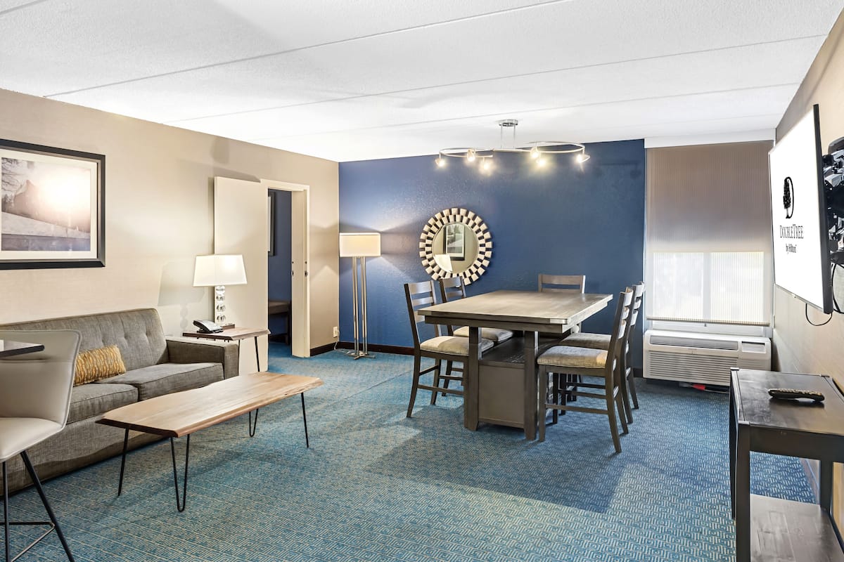 A hotel room with a blue accent wall, a dining table with chairs, a sofa, a coffee table, a TV, and a framed picture on the wall.