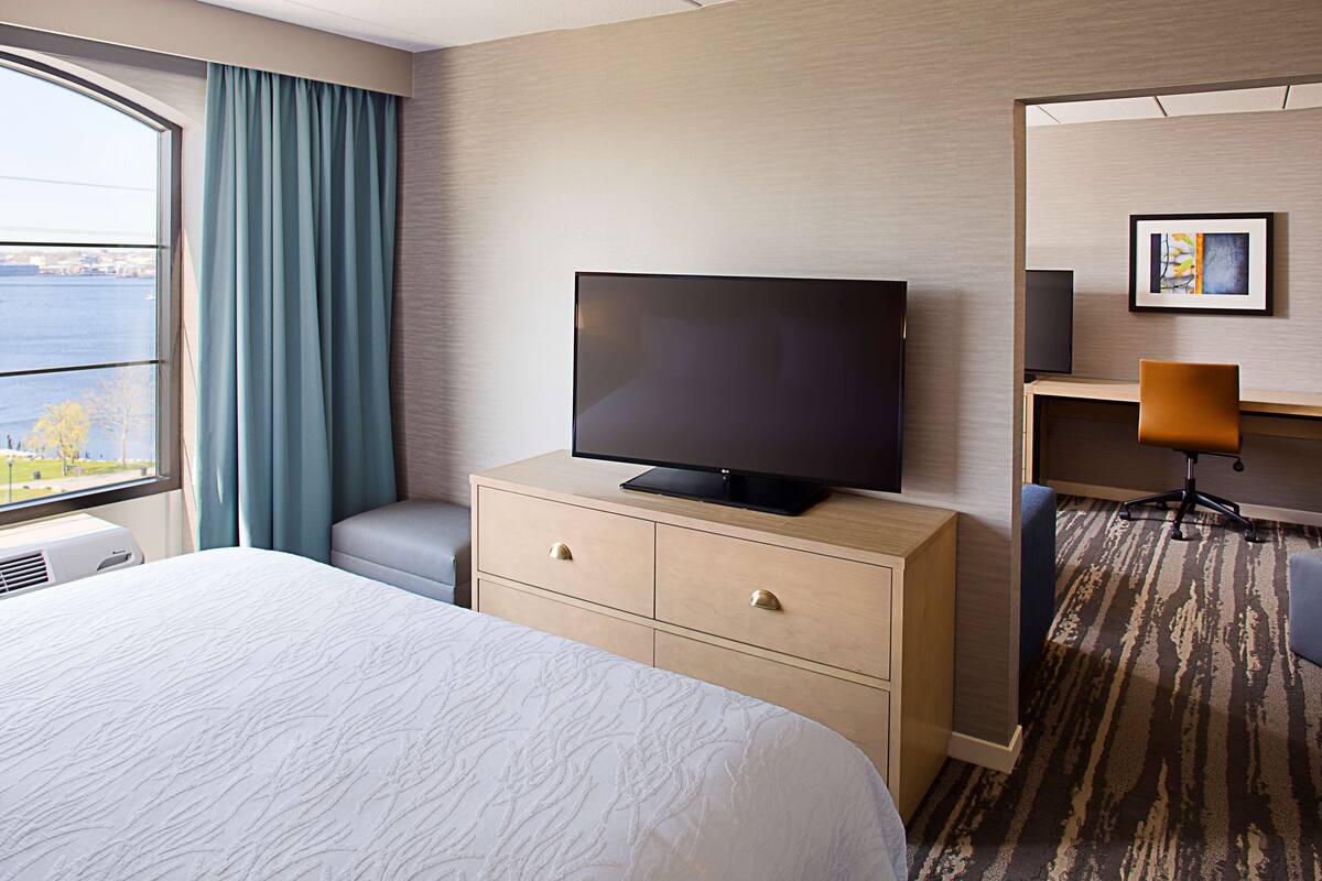 A hotel room with a large bed, a television on a wooden dresser, a desk with a chair, and a view of the water outside.