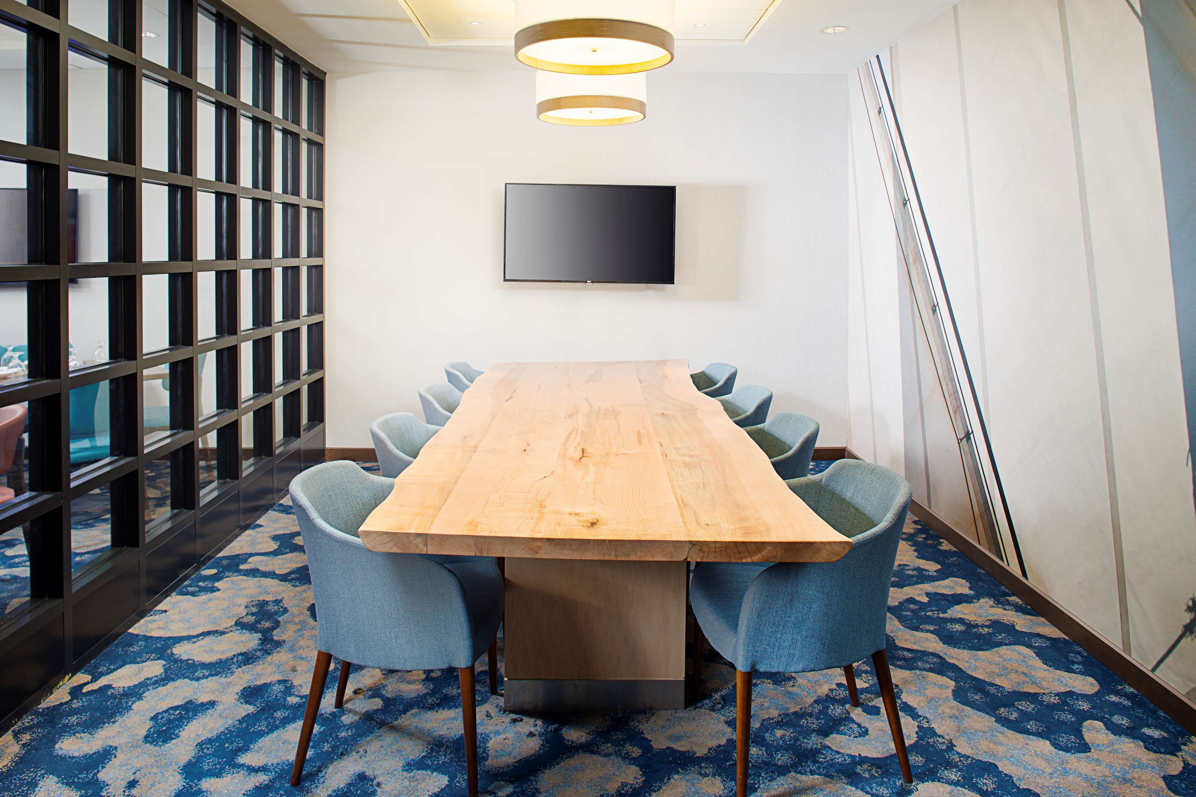 A conference room with a long wooden table, six blue chairs, a flat-screen TV on the wall, and a patterned carpet.