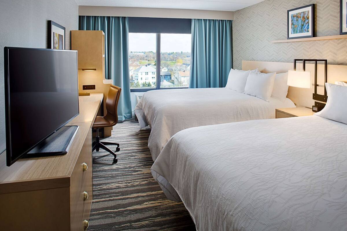 A hotel room with two beds, a desk with a TV, a chair, and a view of the outside.