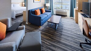 A hotel room with a bed, a blue sofa with orange pillows, a grey armchair, a small table, a TV, and a large window with a view of the outdoors. - Hilton Garden Inn Providence (Providence)