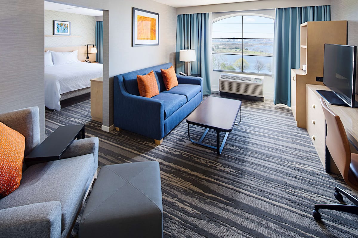 A hotel room with a bed, a blue sofa with orange pillows, a grey armchair, a small table, a TV, and a large window with a view of the outdoors.