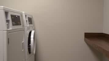 A laundry room with a front-loading washer and dryer, a wooden shelf, and a light-colored floor.