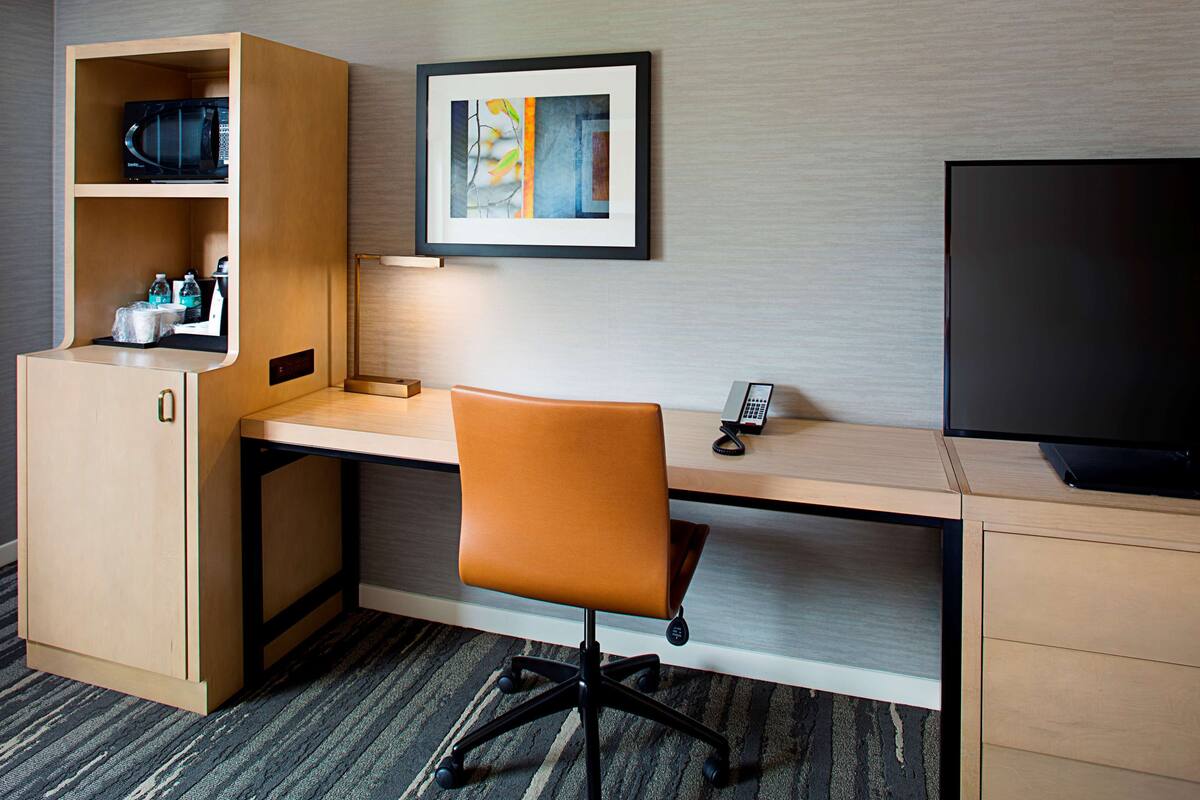 A hotel room with a wooden desk, an orange office chair, a flat-screen TV, a microwave, a coffee maker, and a framed picture on the wall.