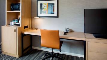 Habitación de hotel con un escritorio de madera, una silla de oficina naranja, un televisor de pantalla plana, un microondas, una cafetera y un cuadro en la pared.