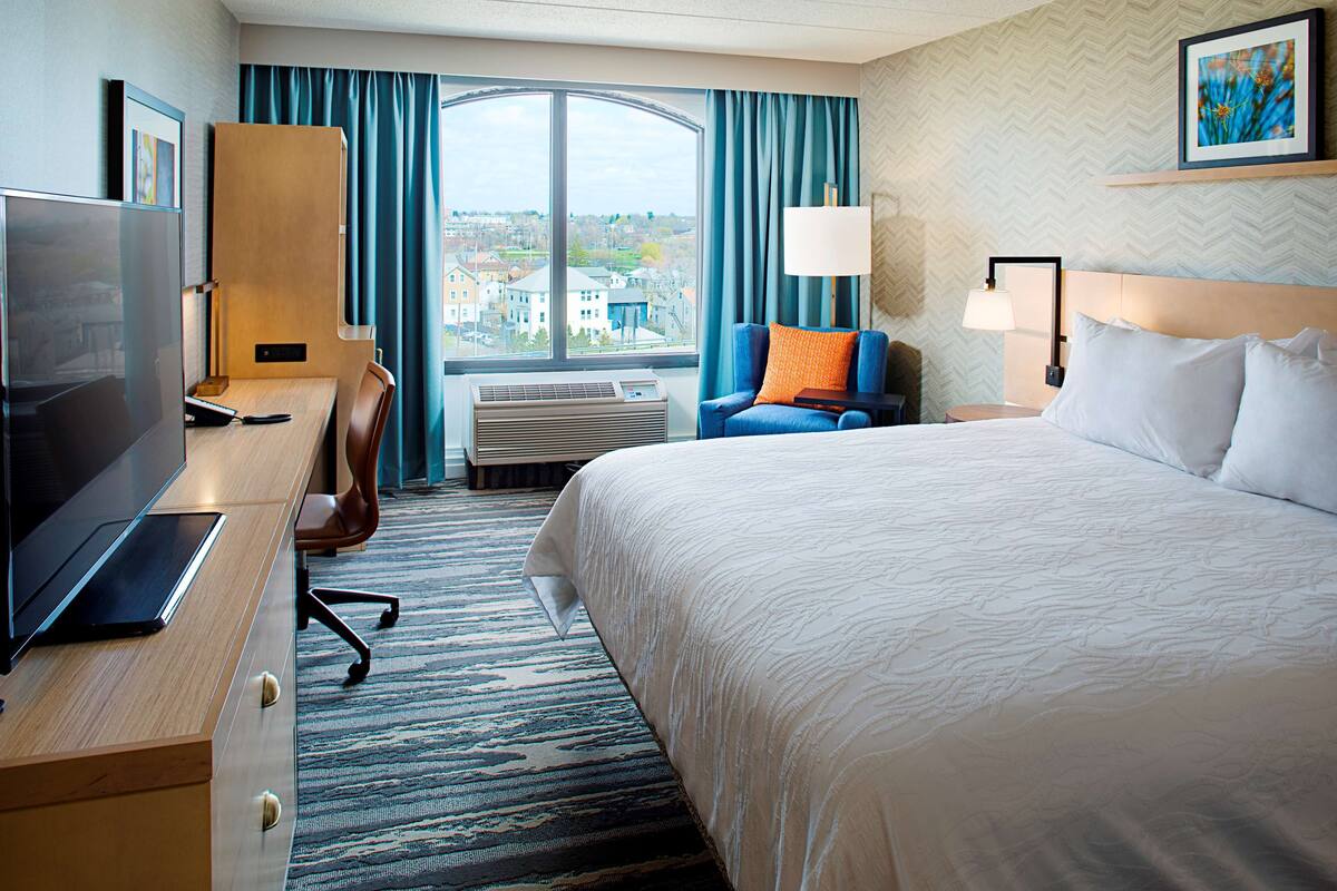 A hotel room with a large bed, a desk with a TV, a chair, a window with curtains, and a city view.