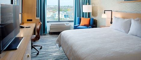 Ein Hotelzimmer mit einem großen Bett, einem Schreibtisch mit Fernseher, einem Sessel, einem Fenster mit Vorhängen und Blick auf die Stadt.