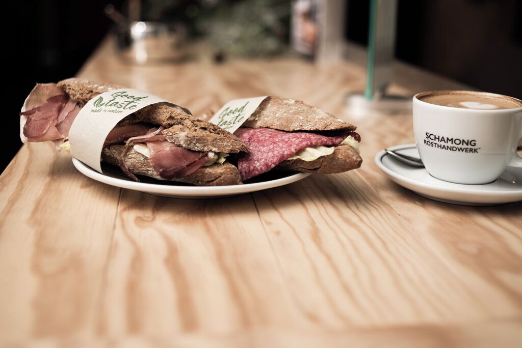 A sandwich wrapped in paper labeled "Good Taste" on a wooden table, next to a cup of coffee with "SCHAMONG RÖSTHANDWERK" branding.