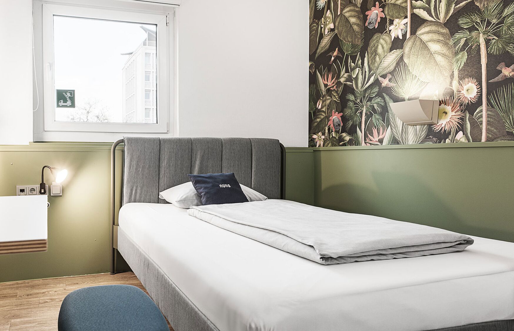 Un lit bien fait, avec une tête de lit grise, une lampe de chevet fixée au mur et un mur vert orné d’un papier peint à motifs tropicaux.