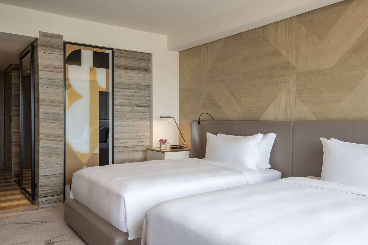 Una habitación de hotel moderna con dos camas, una pared decorativa de madera, una mesita de noche con lámpara y una puerta corrediza que da a un pasillo.