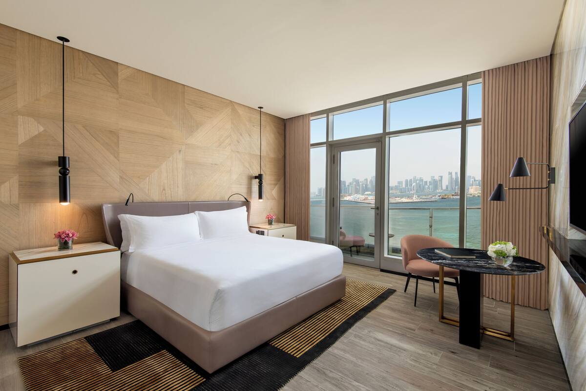 Una habitación de hotel moderna con una cama grande, un escritorio y vistas a un horizonte urbano a través de amplios ventanales.
