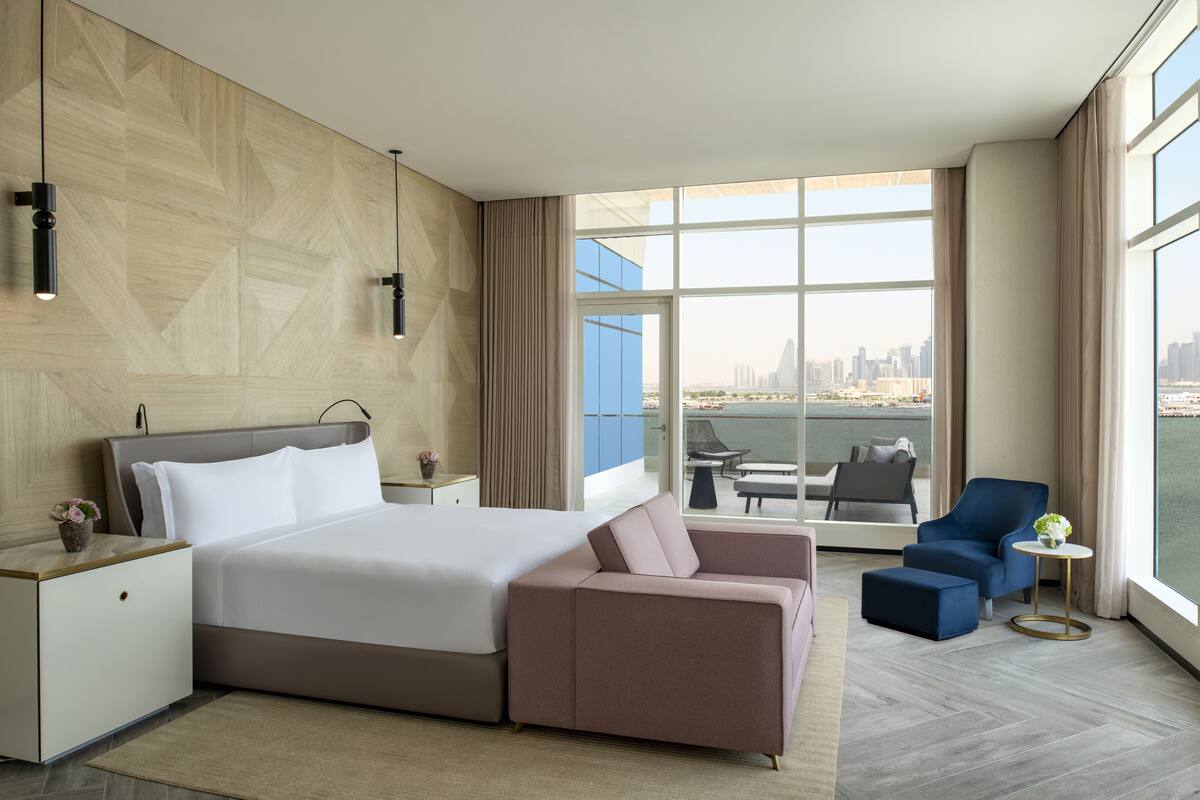 Una habitación de hotel moderna con una cama grande, un sofá, una mesita y vistas a un horizonte urbano a través de amplias ventanas.