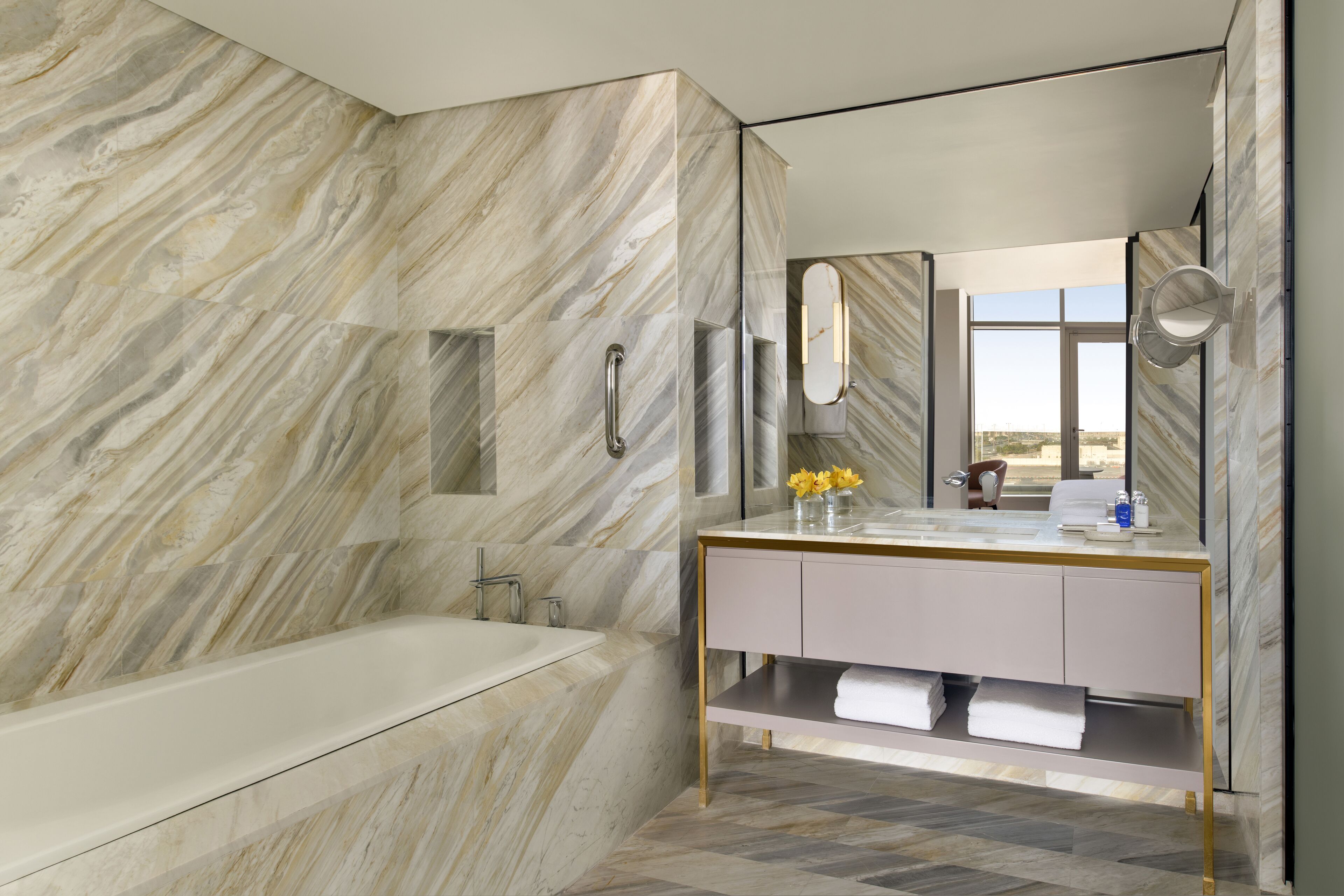 Un baño moderno con pared de mármol, una bañera, un tocador con espejo y una ventana con vistas.