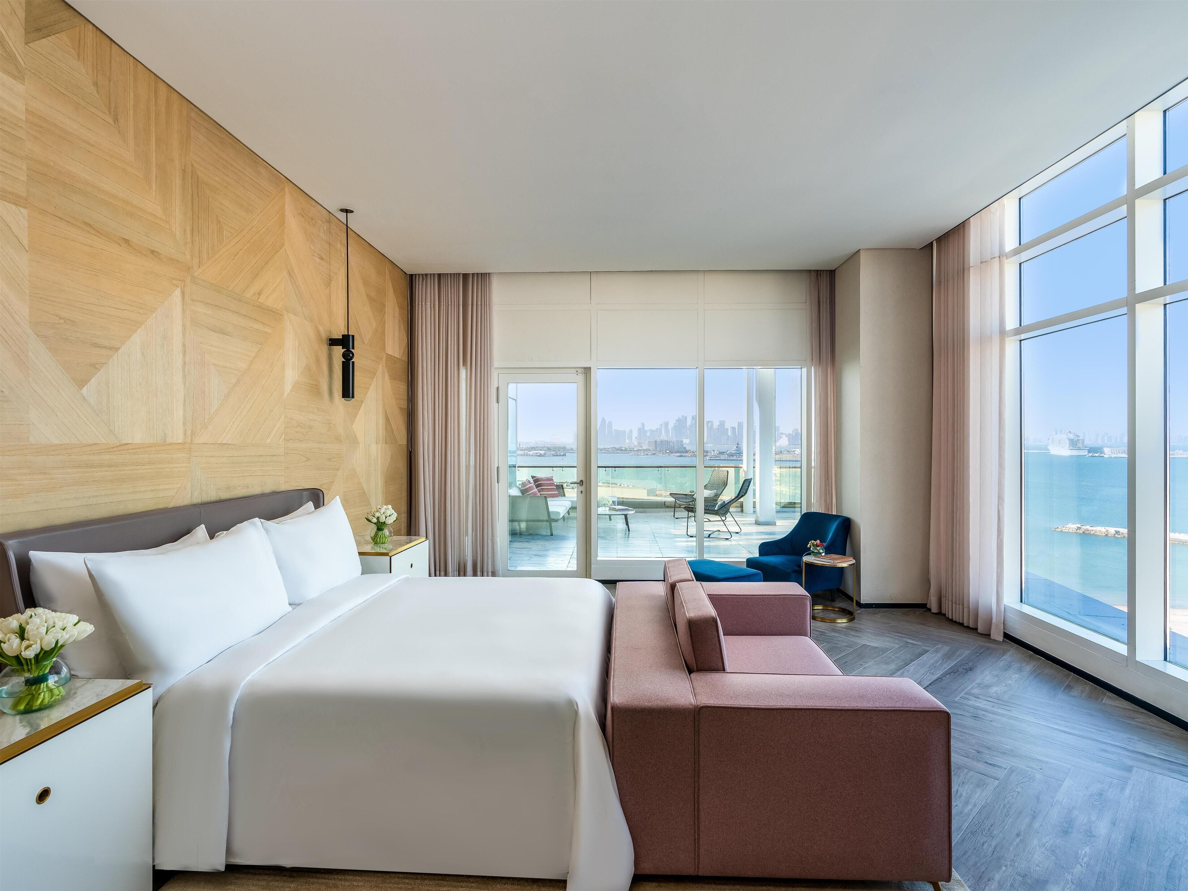 A modern hotel room with a large bed, a seating area with a sofa and armchairs, and a balcony with a city view.