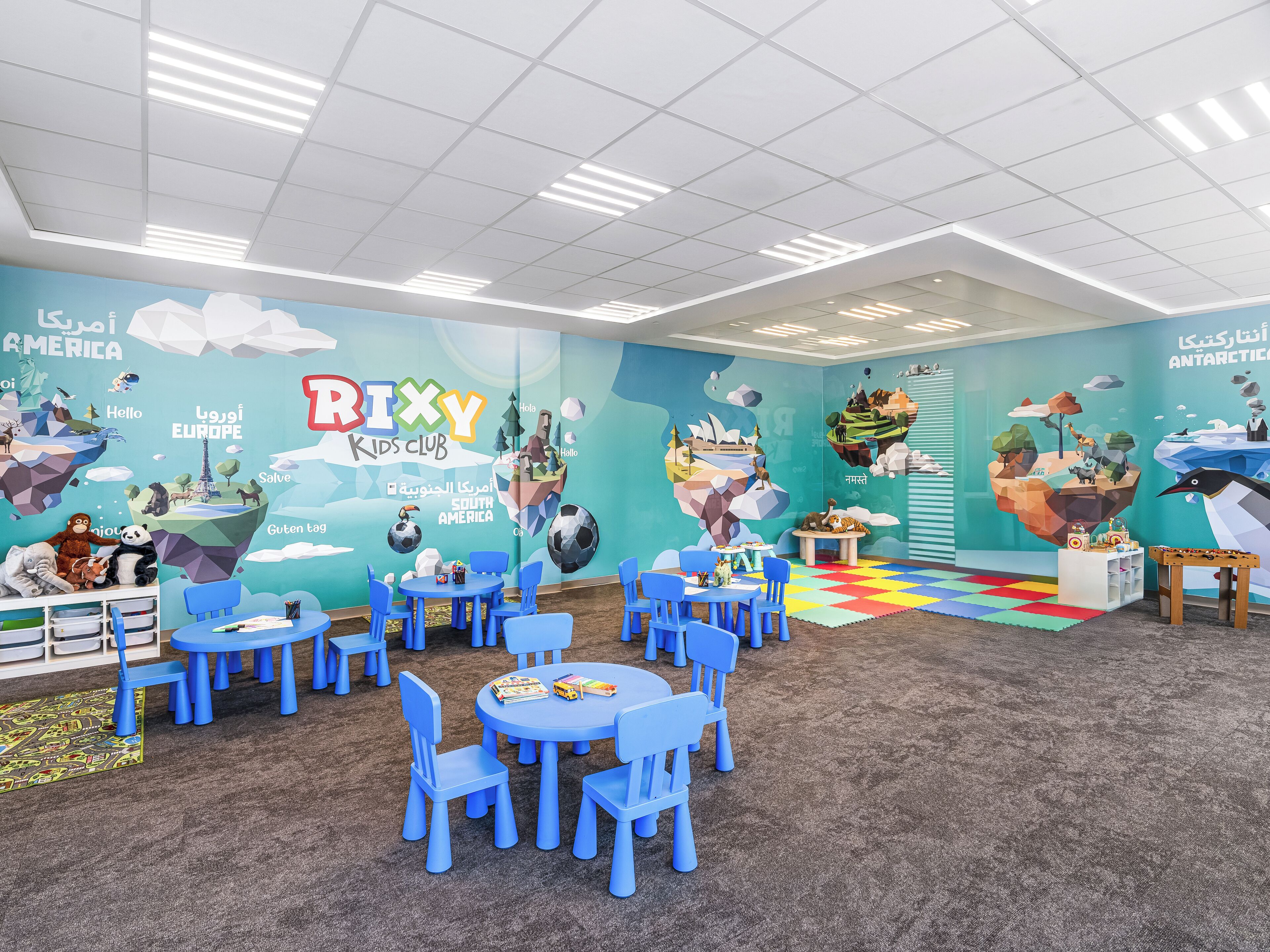 Un espace de jeux pour enfants avec des tables rondes et des chaises bleues, agrémenté d’une fresque colorée représentant les continents et les animaux.