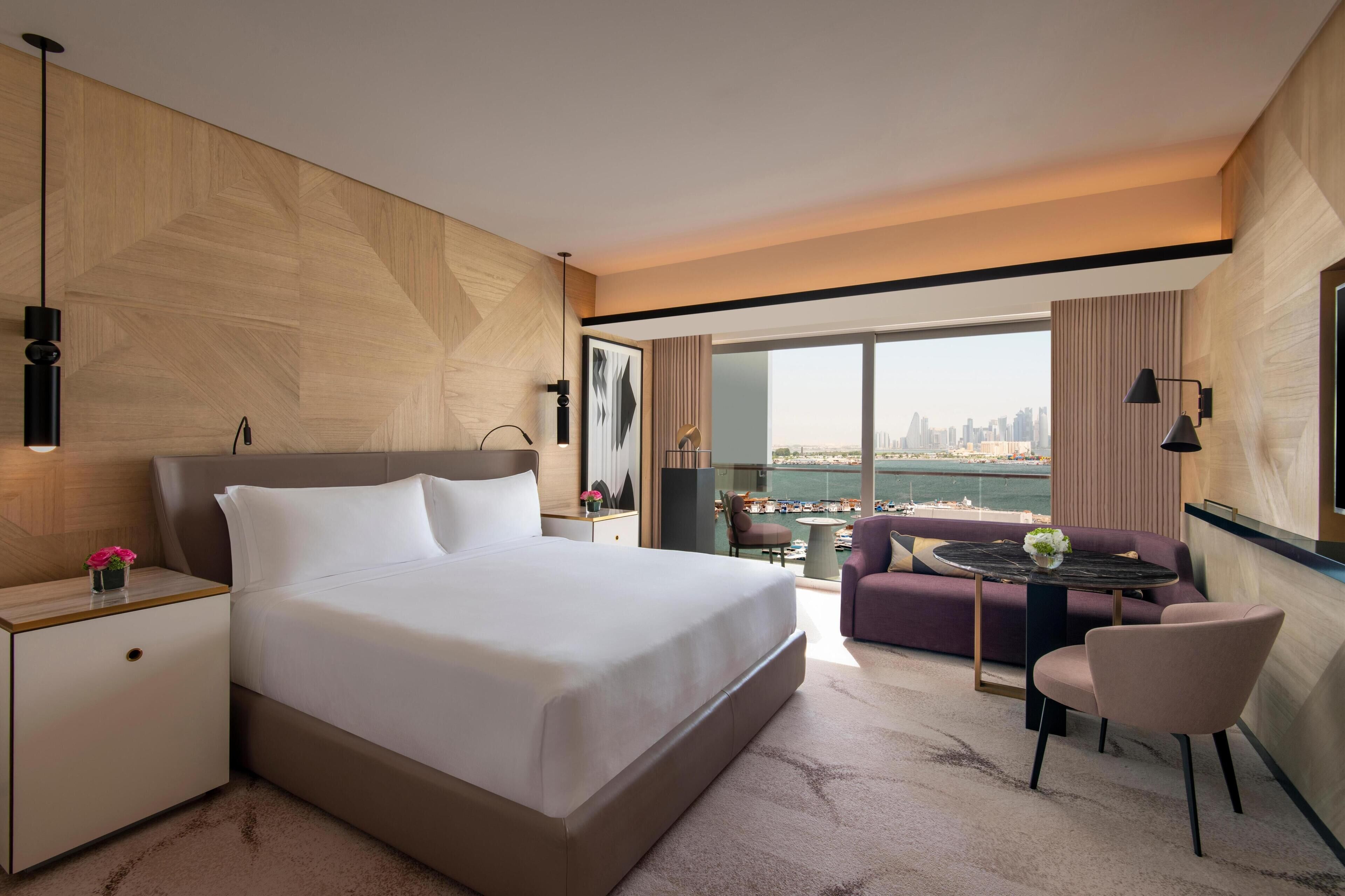 A modern hotel room with a large bed, a small table with a vase, a sofa, and a view of the city skyline.
