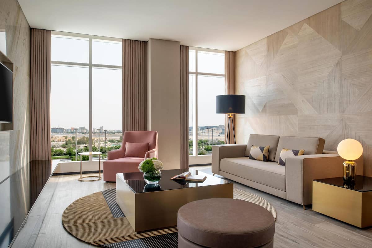 Una habitación de hotel moderna con un ventanal grande, un sofá, una silla, una mesa de centro y una lámpara.