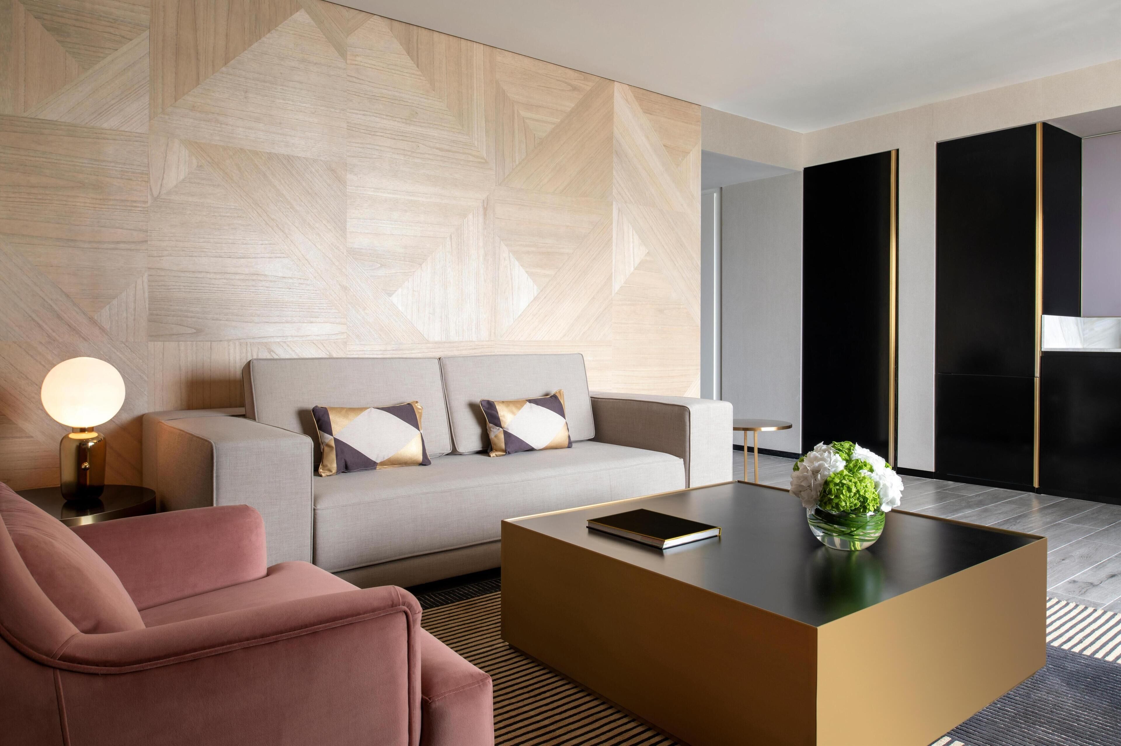 A modern hotel room with a sofa, a pink armchair, a coffee table with a book and flowers, and a geometric-patterned wall.