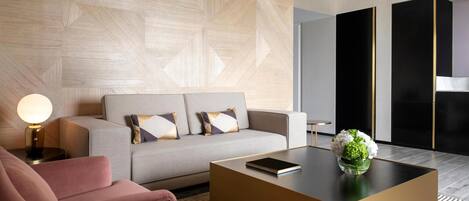 A modern hotel room with a sofa, a pink armchair, a coffee table with a book and flowers, and a geometric-patterned wall.