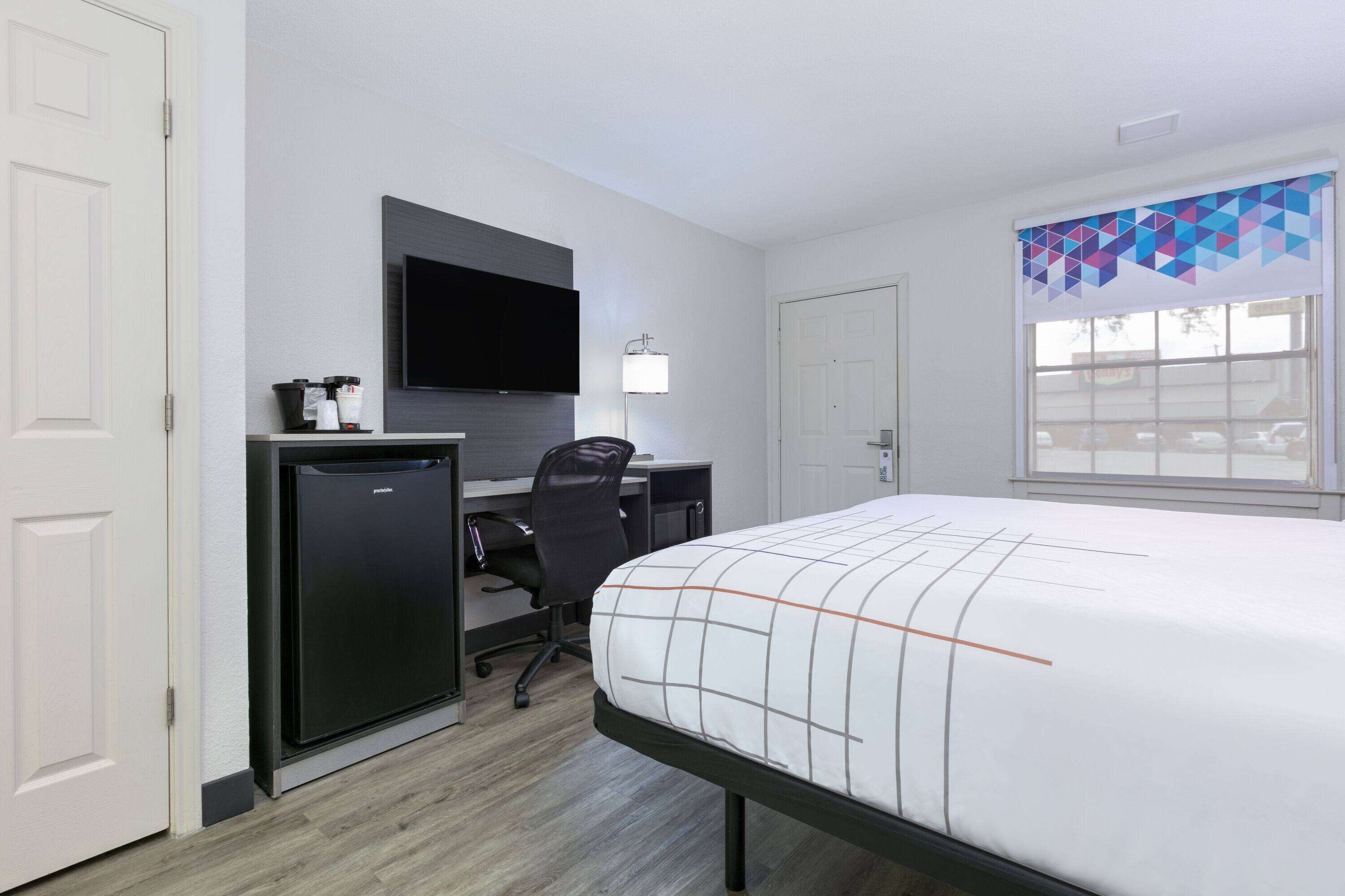 A hotel room with a bed, a desk with a computer, a refrigerator, a TV, and a window with a geometric curtain.