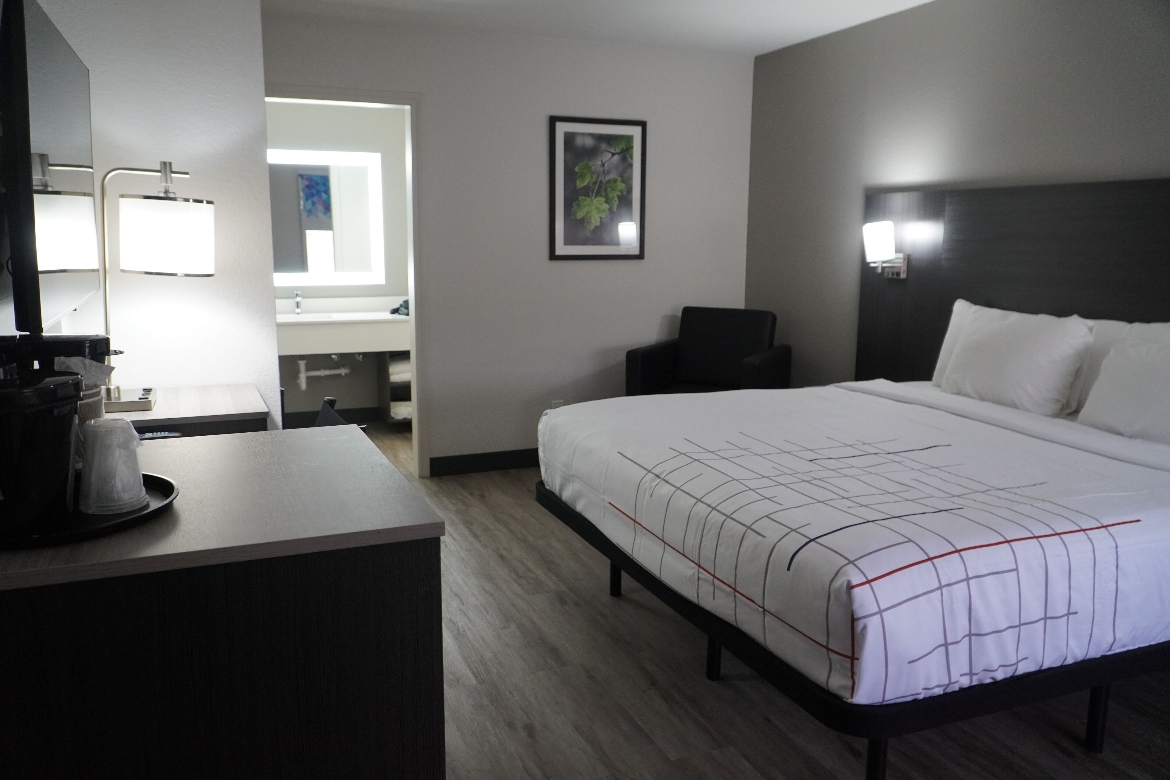A hotel room with a large bed, a desk with a coffee maker, a chair, a framed picture on the wall, and a bathroom visible through an open door.