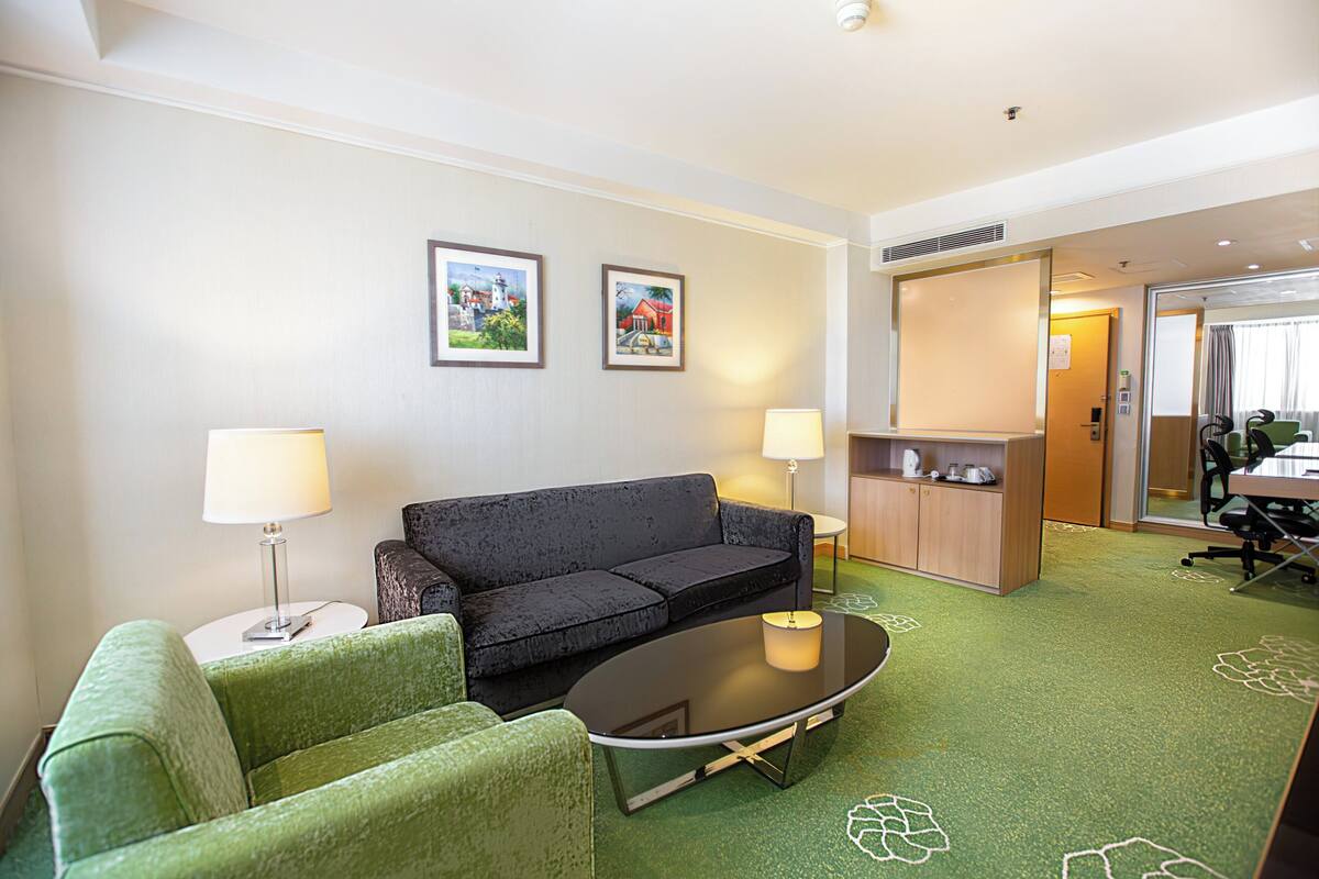 A hotel room with a green sofa, a black sofa, a small round table, two lamps, and a cabinet with a mirror.